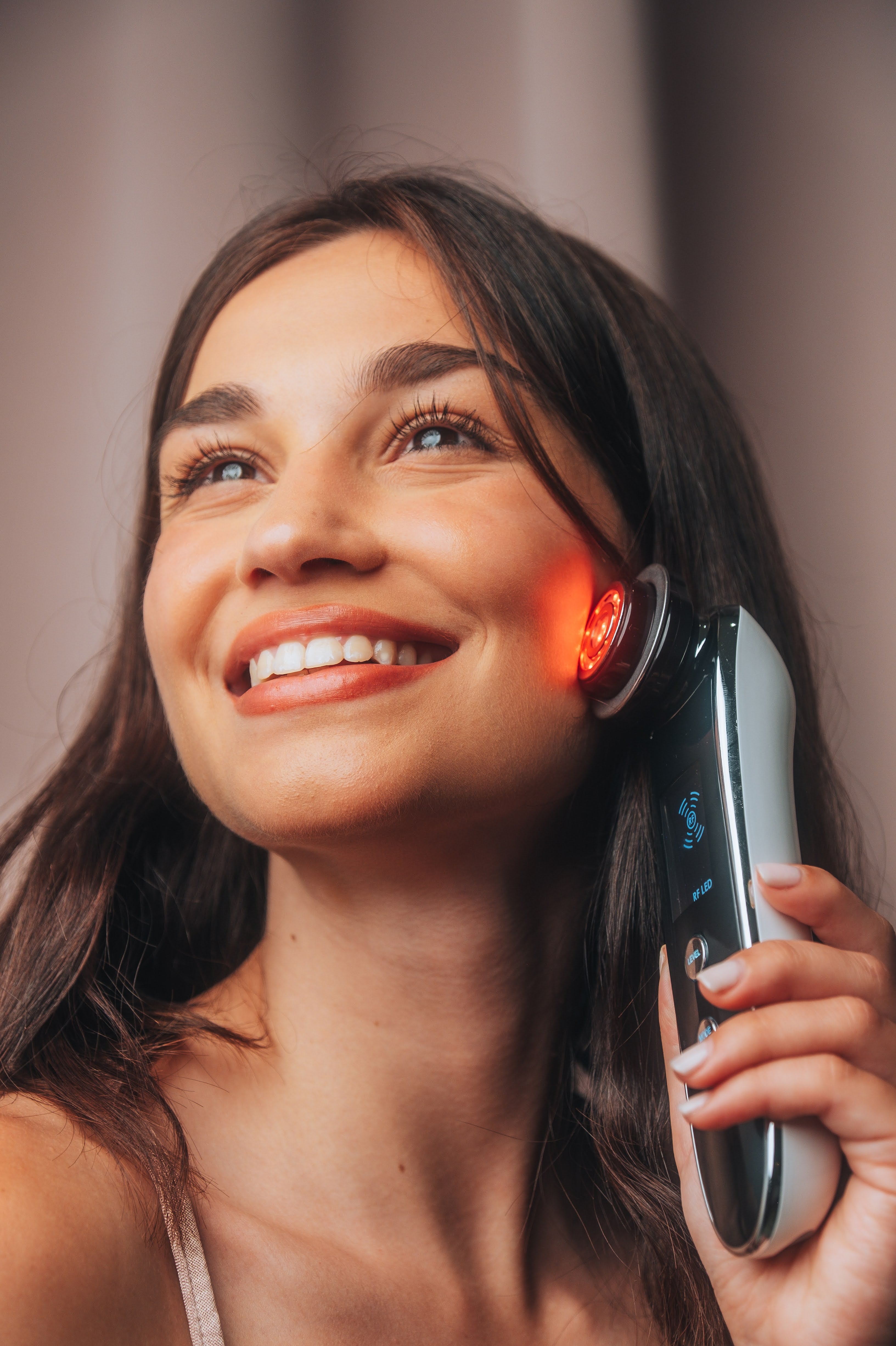 Smiling woman using Fav Naomi Cocoon RF skin-tightening wand on cheek with warm red light